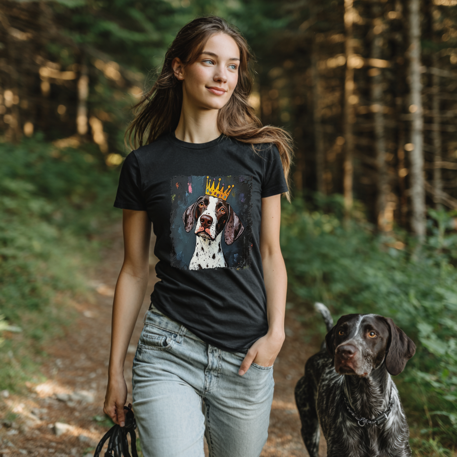 Woman walking in the forest wearing a black Crowned & Connected T-shirt featuring a crowned German Shorthair Pointer dog portrait on the front, with her real German Shorthair Pointer by her side.