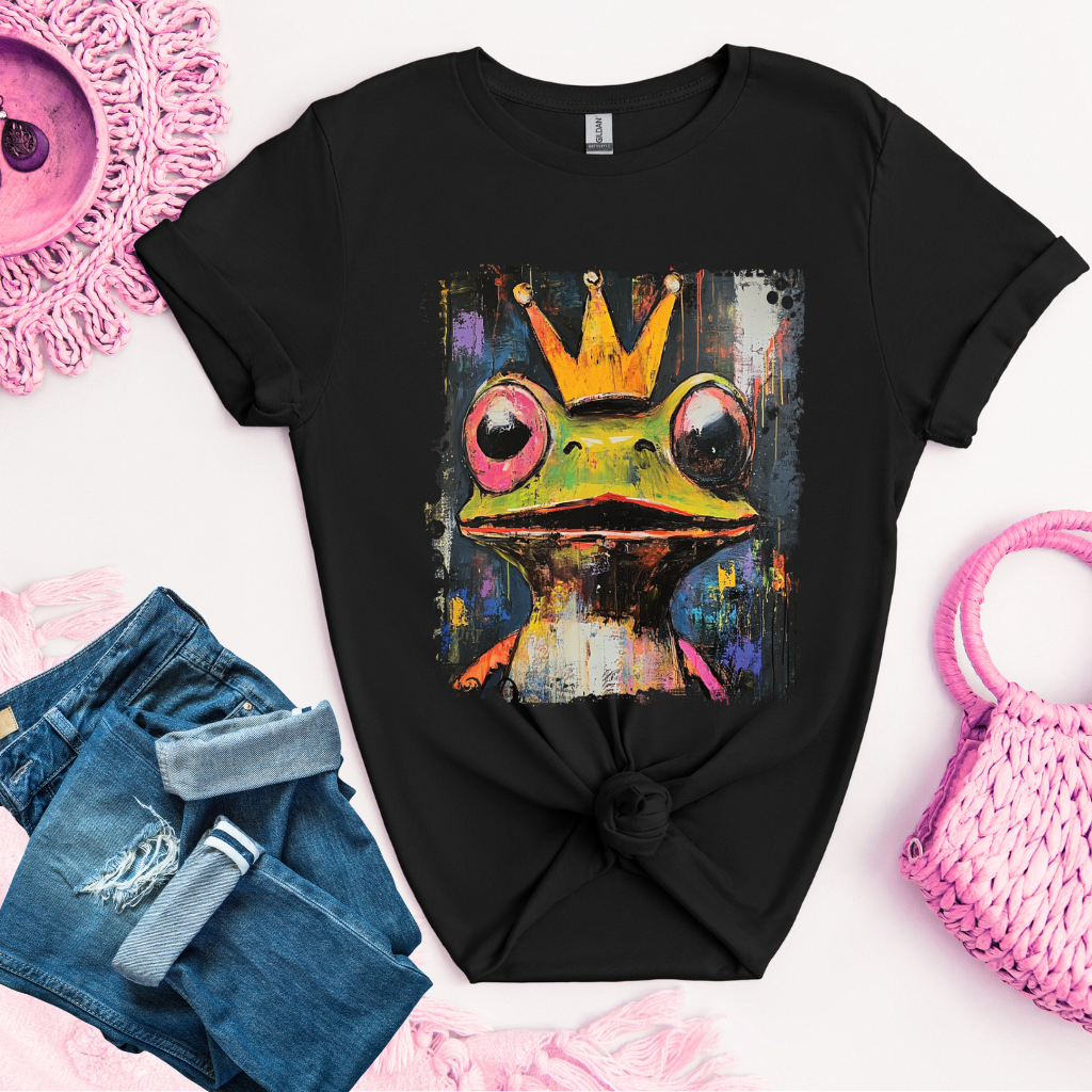 Black t-shirt with a colorful frog wearing a crown graphic, placed on a white surface with denim shorts and a pink bag.