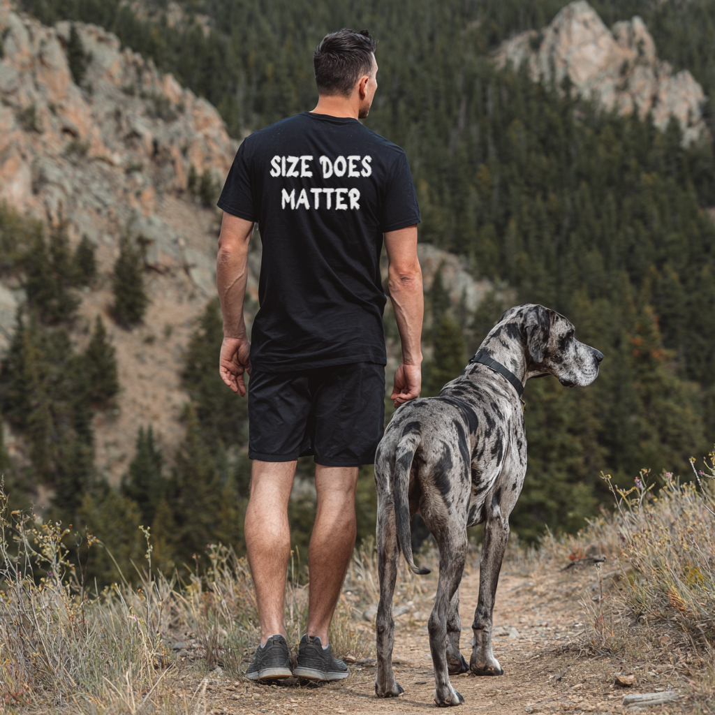 Man wearing a black t-shirt with 'SIZE DOES MATTER' text, walking with a large dog in a mountainous area.