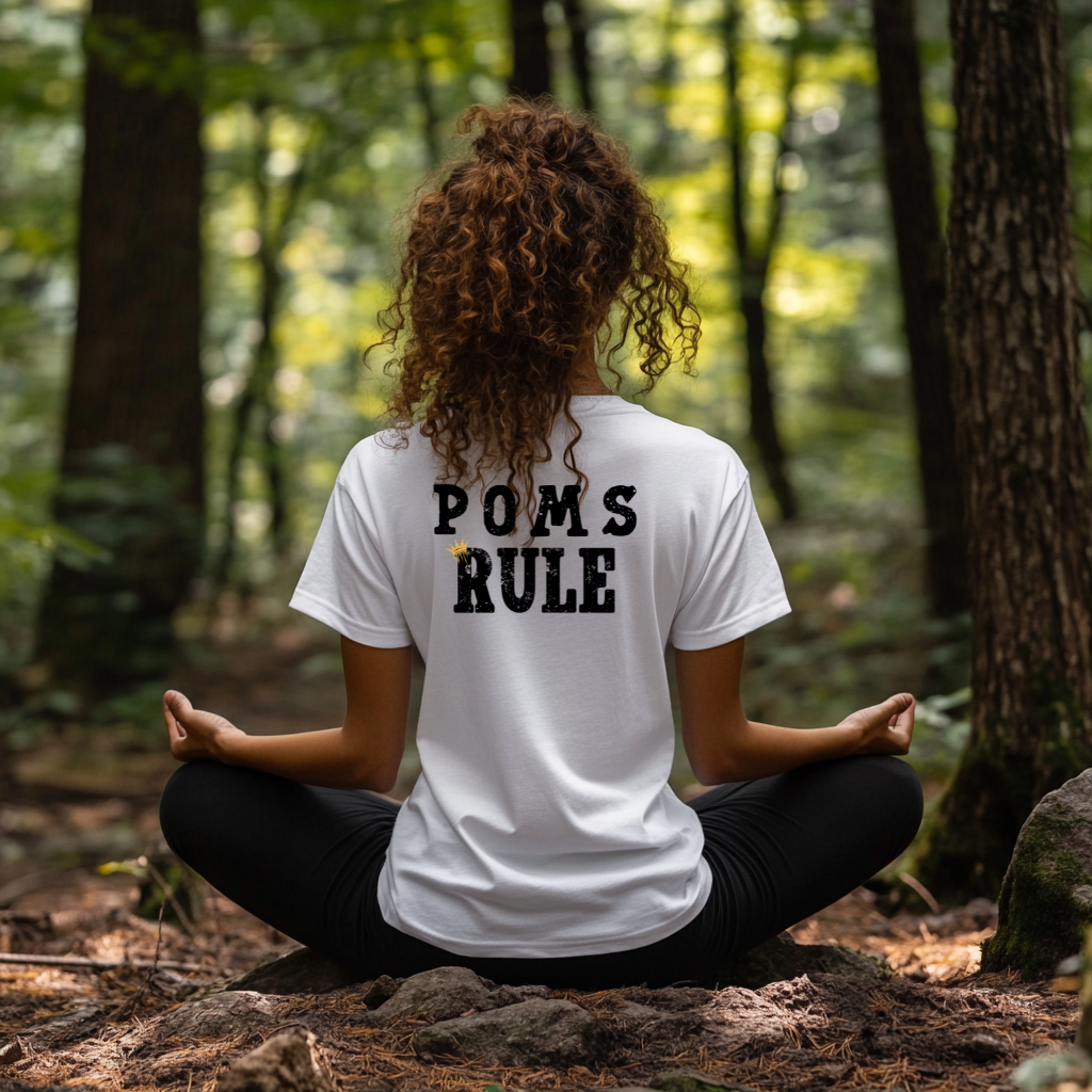 Person sitting in a meditative pose in a forest wearing a white t-shirt with 'POMS RULE' text on the back.