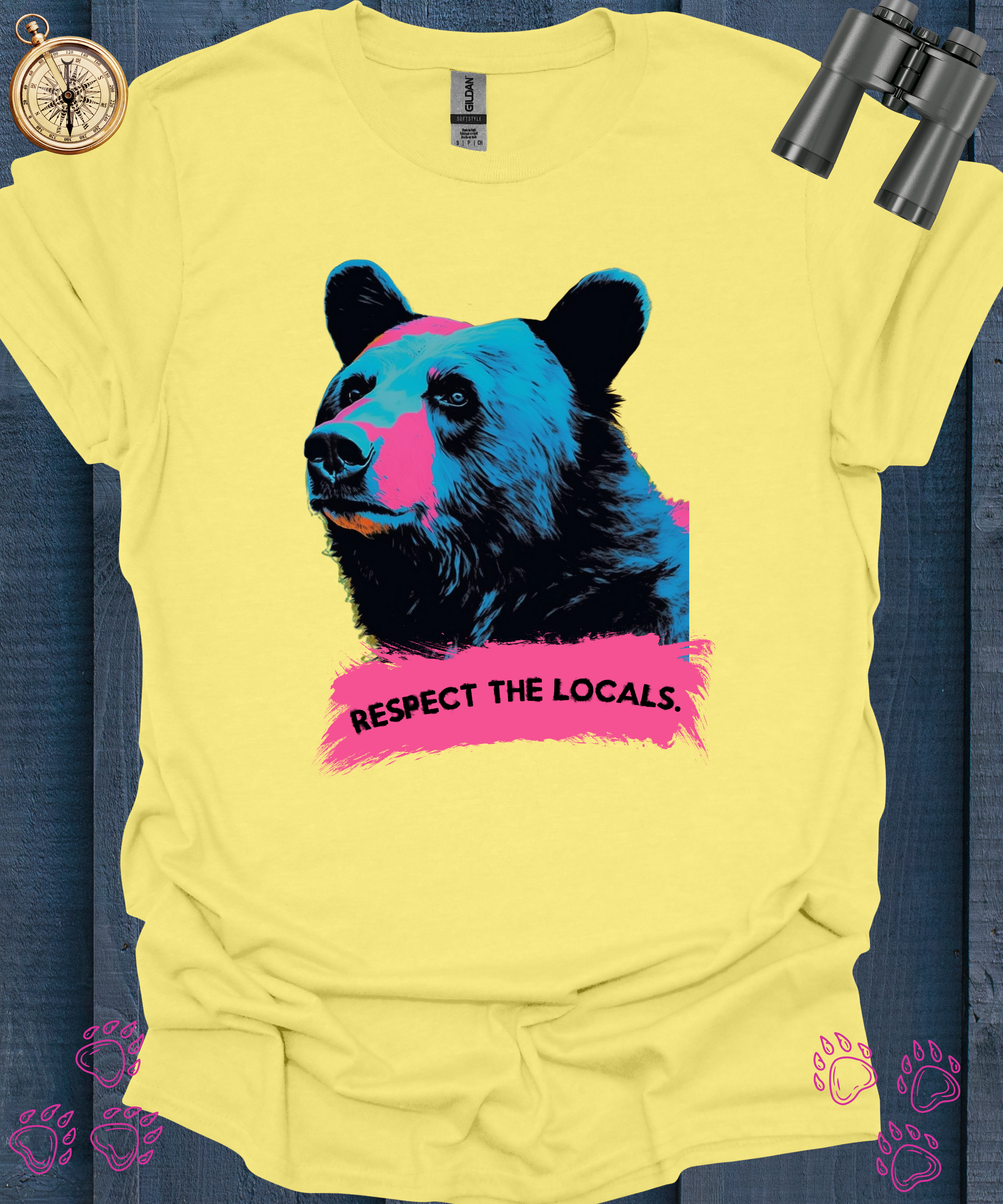 Cornsilk yellow t-shirt featuring a colorful, stylized bear illustration with vibrant blue and pink hues, accompanied by the text 'RESPECT THE LOCALS' in bold pink lettering at the bottom. The t-shirt is displayed on a textured blue wooden background, surrounded by binoculars and toast with peanut butter and jelly, with pink bear paw prints accentuating the scene